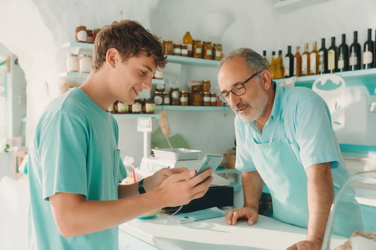 A member shows his phone to a shopkeeper across the counter.